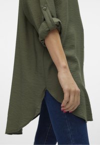 Camicia a maniche lunghe verde oliva con maniche arrotolabili, orlo curvo e tessuto texturizzato, abbinata a jeans blu.