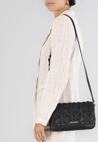 Černá kožená kabelka s tkanou texturou a popruhem, s kovovým logem jako akcentem. Kompaktní obdélníkový tvar, navržená pro nošení přes tělo.