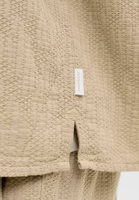 Tissu beige texturé avec une petite fente latérale et une étiquette blanche de la marque Jack & Jones attachée près de l'ourlet.