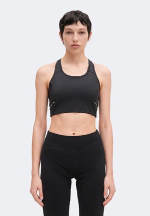 Kvinde med kort mørkt hår iført en sort cropped tanktop og højtaljede sorte leggings, stående mod en ensartet lys baggrund.