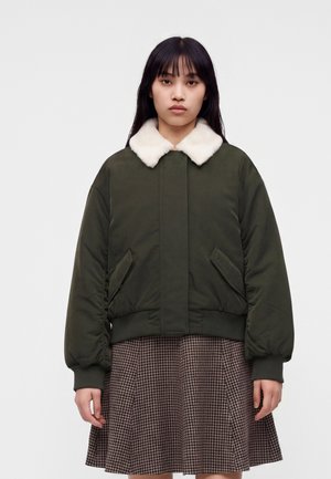 Jeune femme aux cheveux raides et foncés portant une veste vert foncé avec un col en fourrure blanche et une jupe marron à carreaux, debout devant un fond blanc.