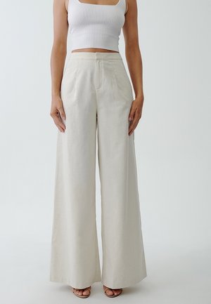 Femme portant un pantalon beige taille haute, jambes larges avec plis à l'avant et un top court blanc côtelé, debout pieds nus sur un fond uni.