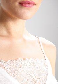 Camiseta blanca con un intrincado borde de encaje en el escote, fabricada con un tejido suave y con finas tirantes ajustables que resaltan la zona del pecho.