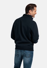 Marineblauwe fleecetrekker met een hoge kraag, gestructureerde stof, ribgebreide manchetten en zoom, gecombineerd met vervaagde blauwe jeans, gezien vanuit de achterkant.