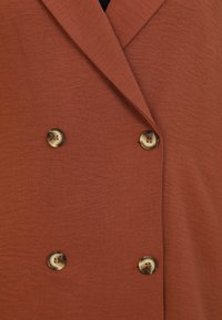 Veste double boutonnage couleur rouille avec une finition texturée. Équipée de grands boutons ronds à motifs sur le devant.