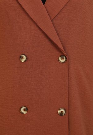Veste double boutonnage couleur rouille avec une finition texturée. Équipée de grands boutons ronds à motifs sur le devant.