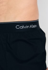 Črne bombažne boksarske hlačke z gladko teksturo, ki imajo na pasu logo Calvin Klein v belem tisku.