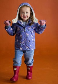 Lila Kinderregenjacke mit Einhornmotiv, weiße fleecegefütterte Kapuze, hellblaue Cordhosen und rote Gummistiefel.