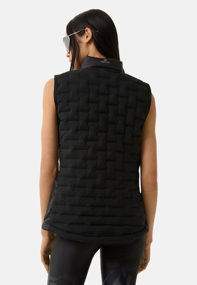 Schwarze ärmellose gesteppte Weste mit hohem Kragen, das eine strukturierte Musterung und ein dezentes Markenlogo auf dem Rücken aufweist. Über dunklen Leggings getragen.