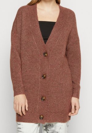 Kvinde iført en lang, brun cardigan med knapper over en sort top og hvide batikfarvede bukser, stående mod en ensfarvet baggrund.