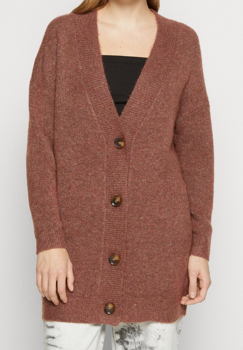 Femme portant un long cardigan marron boutonné sur un haut noir et un pantalon blanc à effet tie-dye, debout devant un fond uni.