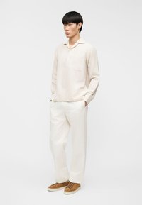 Νεαρός άνδρας με εκρού μακρυμάνικο πουκάμισο, λευκό παντελόνι και καφέ αθλητικά παπούτσια, στέκεται με τα χέρια στις τσέπες μπροστά σε λευκό φόντο.