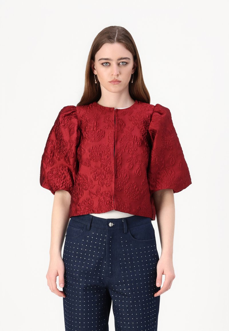 Rote, strukturierte, verkürzte Bluse mit puffärmel, ausgestattet mit einem Reißverschluss an der Vorderseite. Kombiniert mit dunkelblauen Jeans, die mit silbernen Nieten verziert sind.