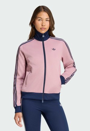 Mujer con una chaqueta deportiva de cremallera rosa y azul marino y leggings azul marino con rayas, de pie con una mano en el bolsillo frente a un fondo liso.