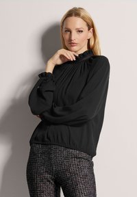 Schwarzes Langarmoberteil mit einem Rüschenausschnitt und gathered cuffs. Glattes Material, lockere Passform; getragen mit gemusterten texturierten Hosen.