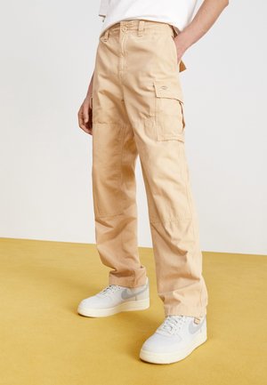 Beige cargobyxor i bomull med lös passform, med flera fickor och sömmar, tillsammans med grå sneakers. Slät yta.
