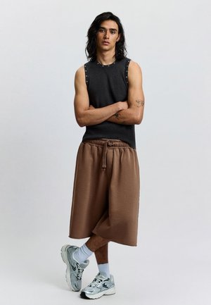 Fiatal felnőtt, vállig érő fekete hajjal, fekete ujjatlan felsőt, barna bőszárú rövidnadrágot, fehér zoknit és ezüst sportcipőt visel, karját összefonva áll.