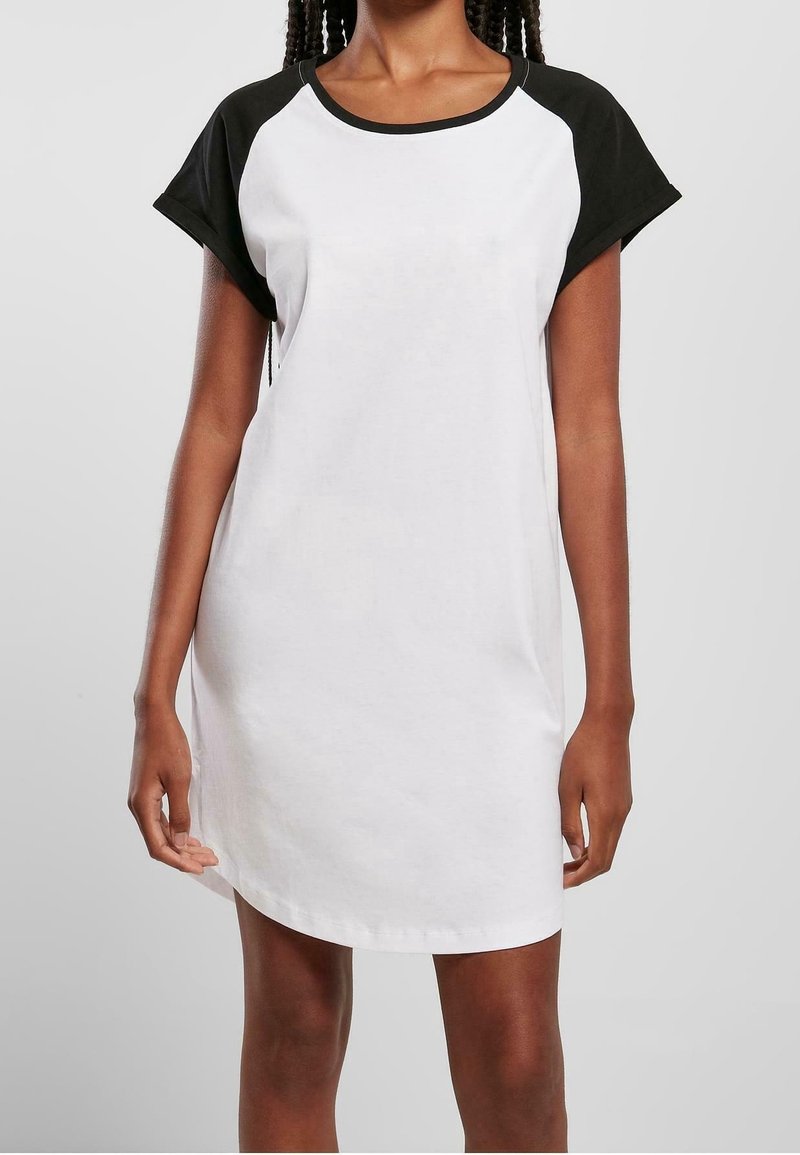 Femme portant une robe blanche à manches courtes, longueur genou, avec des manches raglan noires et un col noir sur un fond clair uni.
