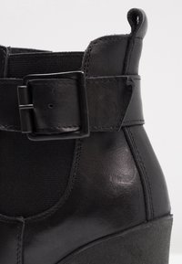 Gros plan sur une botte cheville en cuir noir avec un panneau latéral élastique, une large sangle à boucle carrée et une languette au talon.