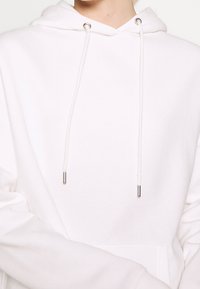 Biała bluza z kapturem ze sznurkiem, srebrnymi metalowymi końcówkami i przednią kieszenią. Materiał wydaje się miękki i lekko teksturowany.