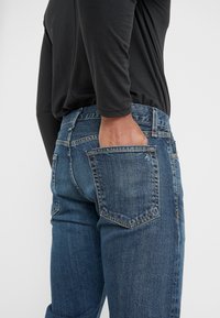 Vaqueros de mezclilla en azul medio con desgaste ligero. Presentan un bolsillo trasero con detalle de costura. Combinados con una blusa negra de manga larga.