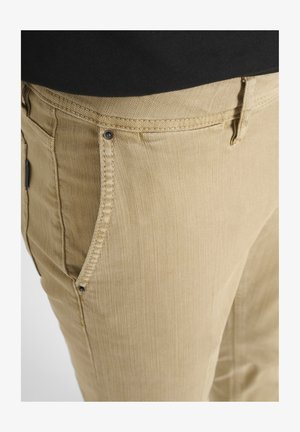Pantaloni in cotone beige con tasche laterali visibili, tessuto texturizzato e dettagli in metallo. I particolari delle cuciture sono evidenti lungo i bordi.
