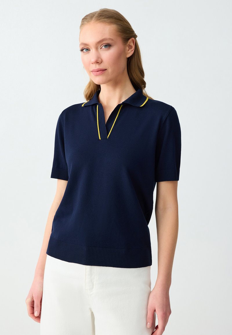 Femme aux cheveux châtain clair portant un polo bleu marine à manches courtes avec un liseré jaune et un pantalon blanc, debout devant un arrière-plan uni clair.