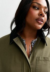 Olivgrön jacka med svart krage, utrustad med knäppning fram och bröstficka. Bärs över en blå och vit rutig skjorta.