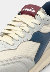Close-up of beige and gray sneaker with cream laces, navy blue side accent, and red tongue label.