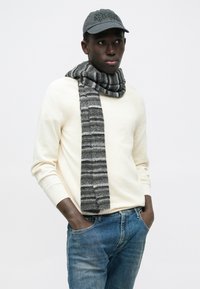 Άνδρας που φοράει γκρι καπέλο, εκρού πουλόβερ, γκρι ριγέ κασκόλ και μπλε τζιν με τα χέρια στις τσέπες, μπροστά σε λευκό απλό φόντο.