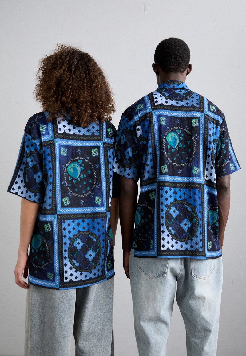 Chemise à manches courtes avec un motif géométrique bleu foncé et noir. Présente des accents de vert et de bleu, une surface texturée et une coupe décontractée.
