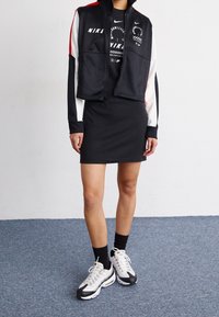 Svart kort jacka med röda och vita accenter, parat med en svart klänning och grå sneakers. Strukturerad tyg, sportig design.