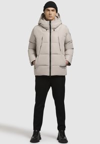 Mann in beiger Steppjacke mit Kapuze, schwarzem Strickmütze, schwarzen Hosen und schwarzen Stiefeln, der mit den Händen in den Taschen vor einem weißen Hintergrund steht.