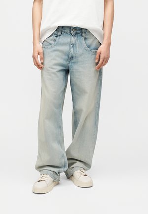 Šviesiai mėlynos laisvo kirpimo džinsai su priekine kišene, dėvimi su baltais sportbačiais ir įkišta balta palaidine ant stovinčio asmens.