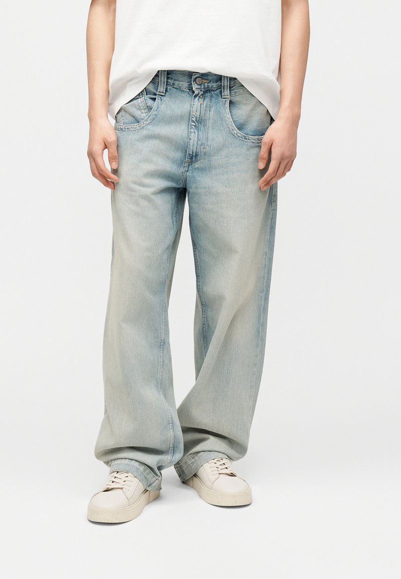 Ανοιχτό μπλε τζιν σε άνετη γραμμή με εμπρόσθιες τσέπες, φορεμένα με λευκά αθλητικά παπούτσια και μία λευκή πουκάμισα μέσα από το παντελόνι, σε ένα πρόσωπο που στέκεται.