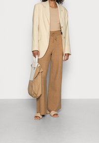Beige blazer over een geribbelde beige top, bruine wijde pantalons met trekkoord, tan geweven sandalen en een lichte beige tote bag.