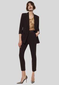 Blazer marrone su misura con design a single-breasted, abbinato a un top con motivo dorato e pantaloni tapered coordinati. Tacchi alti argentati.