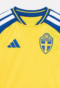 Κίτρινη αθλητική φανέλα με μπλε λεπτομέρειες, που φέρει το έμβλημα της εθνικής ομάδας ποδοσφαίρου της Σουηδίας και τρεις άσπρες λωρίδες στους ώμους.