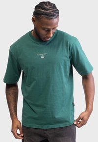 T-shirt en coton vert à manches courtes, col rond côtelé, et petit logo brodé sur la poitrine ; coupe décontractée avec une texture lisse.