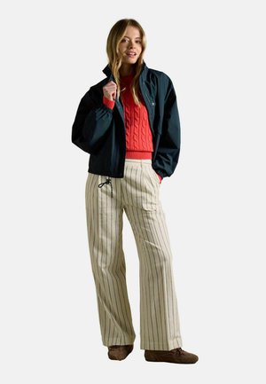 Mladá žena oblečená v červenom svetríku s vrkočovým vzorom, námorníckej bundy, krémových širokých nohaviciach s pruhmi a hnedých topánkach, stojaca s jednou rukou v vrecku.