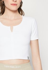 Žena v bílém krátkém rukávu s crop topem a malým výstřihem vpředu, doplněná béžovými kalhotami s logem Calvin Klein Jeans na pase.