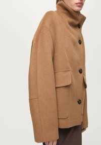 Chaqueta de mezcla de lana color camello con cuello alto, grandes botones y bolsillos laterales. Forma cuadrada, textura suave y acentos de diseño minimalista.