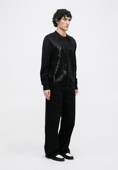 Cárdigan negro de punto con textura acanalada y detalles de lentejuelas, combinado con pantalones negros holgados y zapatos en blanco y negro, sobre un fondo neutro.