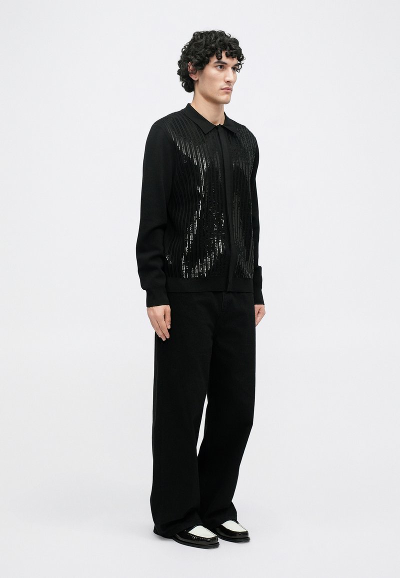 Schwarze Strickjacke mit gerippter Textur und Paillettenverzierung, kombiniert mit lockeren schwarzen Hosen und schwarz-weißen Schuhen, vor einem neutralen Hintergrund.