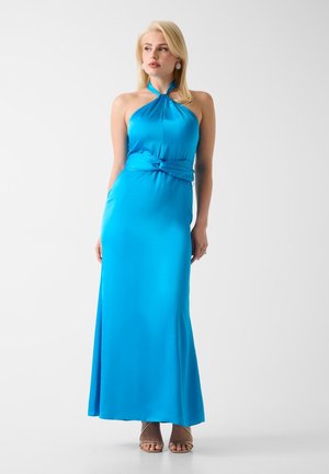 Blonde Frau in einem ärmellosen, blauen Satin-Kleid mit Neckholder und gedrehtem Taillendetail, dazu beige offene Absatzschuhe, vor weißem Hintergrund stehend.