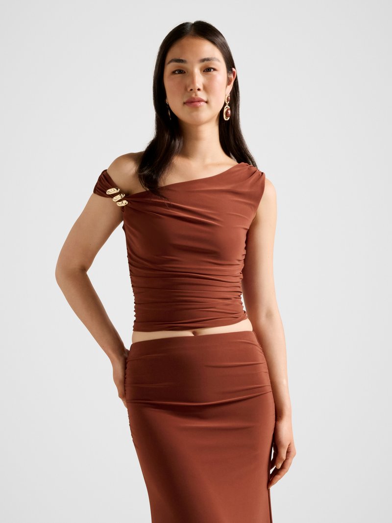 Rjav vrhnji del obleke s potegnjeno tkanino in enostransko obliko ramena; s poudarki v zlati barvi. Ujemajoča se oprijeta rjava krila; gladka, teksturirana tkanina.