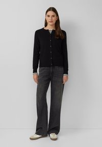 Gilet noir avec un col rond et des boutons à l'avant, associé à un jean gris évasé taille haute. Tissu texturé, détails minimalistes.