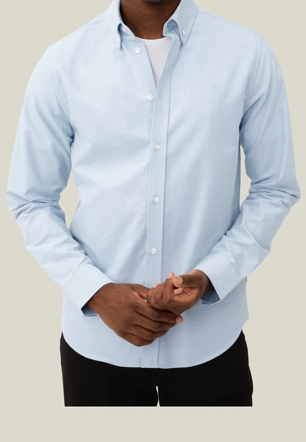 Homme portant une chemise bleu clair boutonnée sur un t-shirt blanc, avec un pantalon noir, debout devant un fond neutre, les mains jointes.