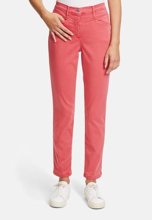 Personne portant un pantalon slim corail rose et des baskets blanches, debout devant un fond clair uni.