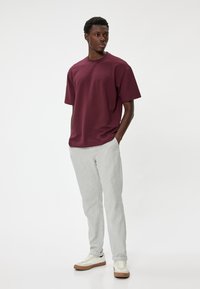 Bordeauxkleurig textuur t-shirt met korte mouwen, gecombineerd met lichtgrijze tapered broek en witte sneakers met bruine zolen. Staande model in profiel.